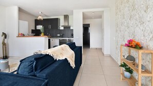 Un salon moderne avec un canapé bleu foncé, une petite table d’appoint en bois ornée de fleurs et, au fond, une cuisine.