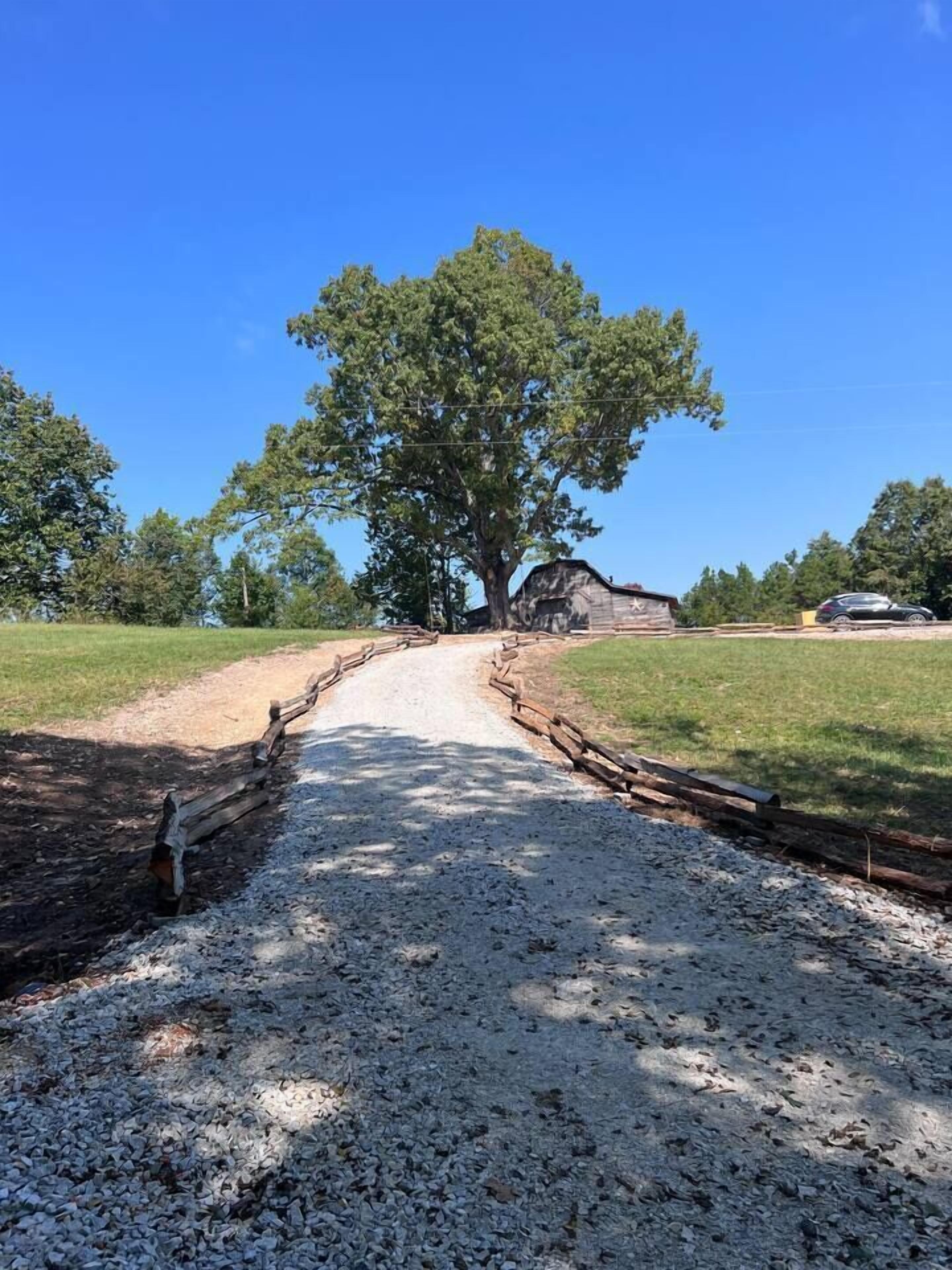 Un chemin de gravier mène à un bâtiment rustique en bois, avec un grand arbre au centre.