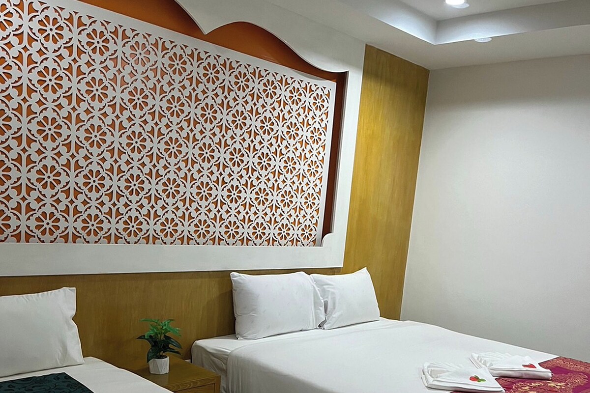 A hotel room with two beds, a decorative wall panel, a small table with a plant, and a red throw blanket.