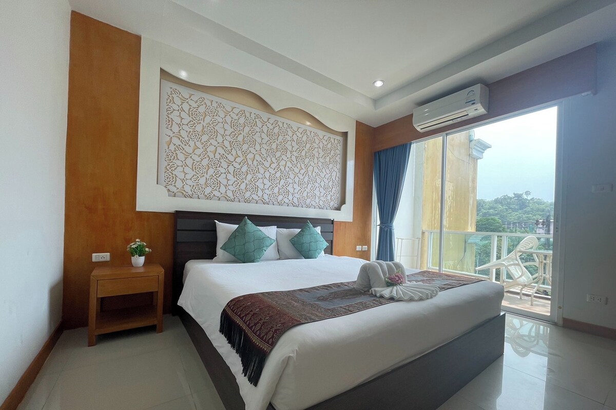 A modern hotel room with a large bed, a decorative wall panel, a small bedside table with a plant, and a balcony with a view.