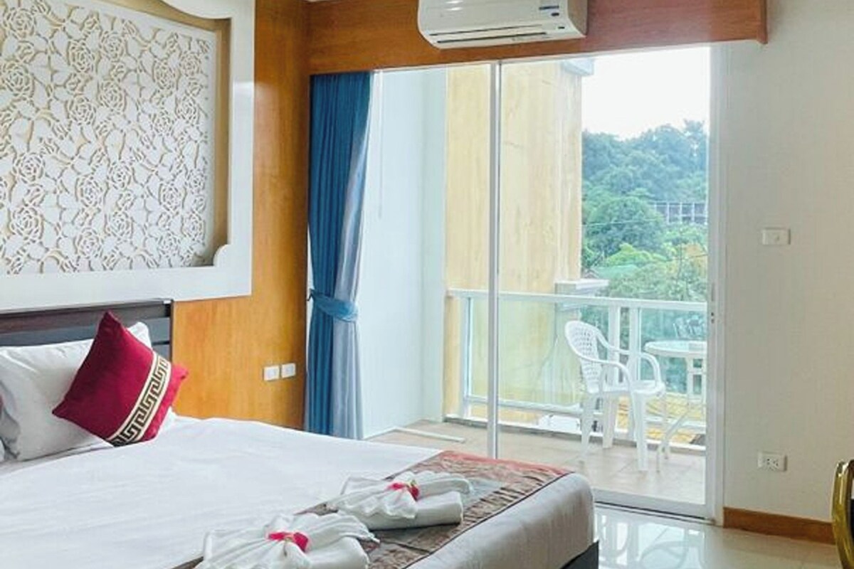 A hotel room with a large bed, a balcony with white chairs, and an air conditioning unit.
