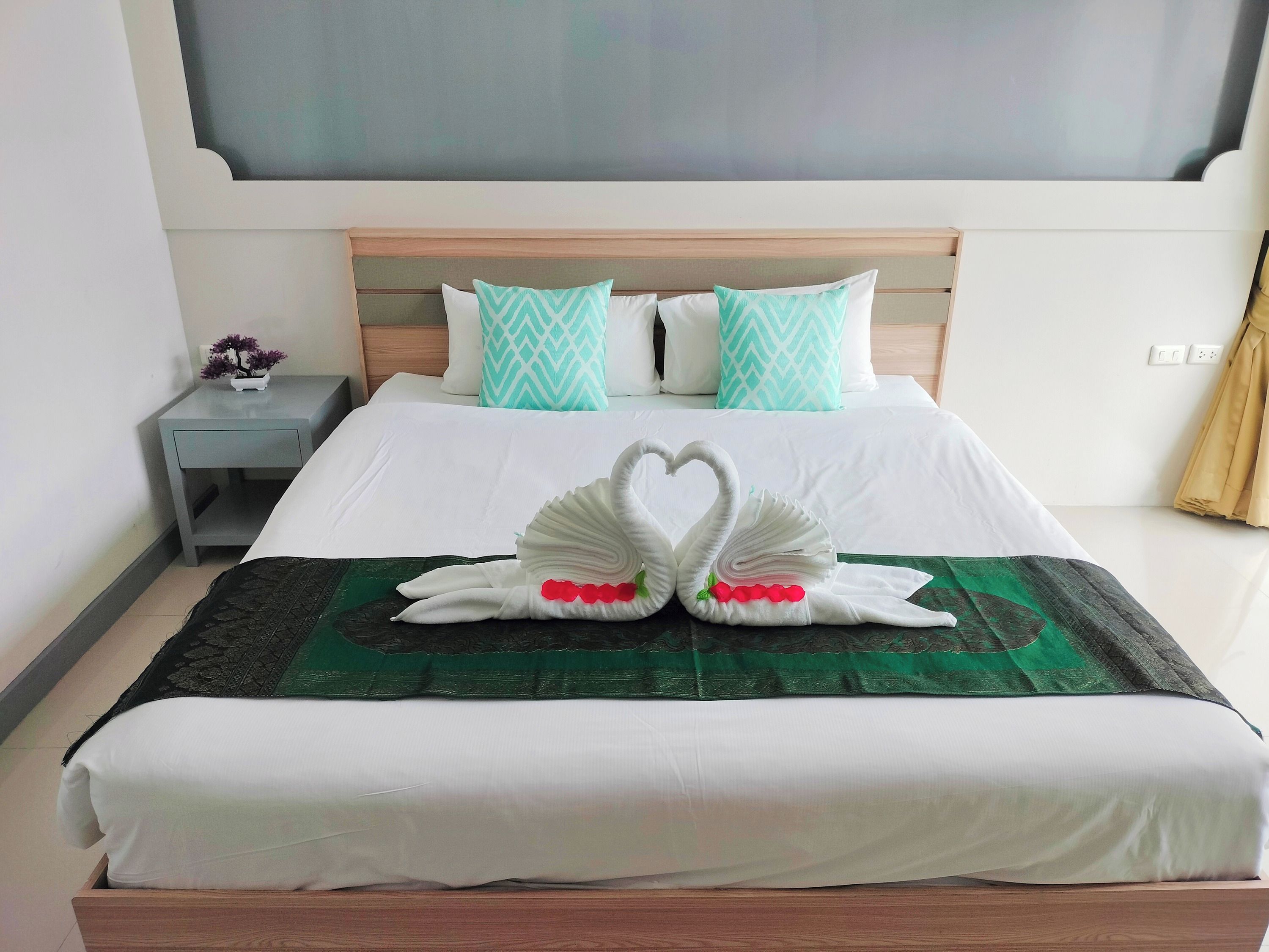 Ein ordentlich gemachtes Bett mit einer grünen Tagesdecke, zwei schwanenförmigen Handtüchern und einem Nachttisch mit einem Blumenstrauß.