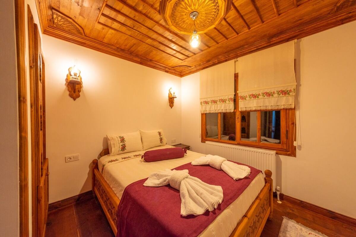 A wooden-paneled bedroom with a bed, two wall sconces, a window with a floral valance, and a radiator.