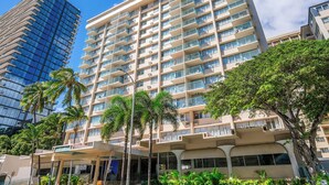 A multi-story hotel with balconies, a modern glass building to the left, and palm trees in the foreground. - Penthouse in Waikiki with ocean & mountain views 5 minutes walk to Waikiki beach (Honolulu)