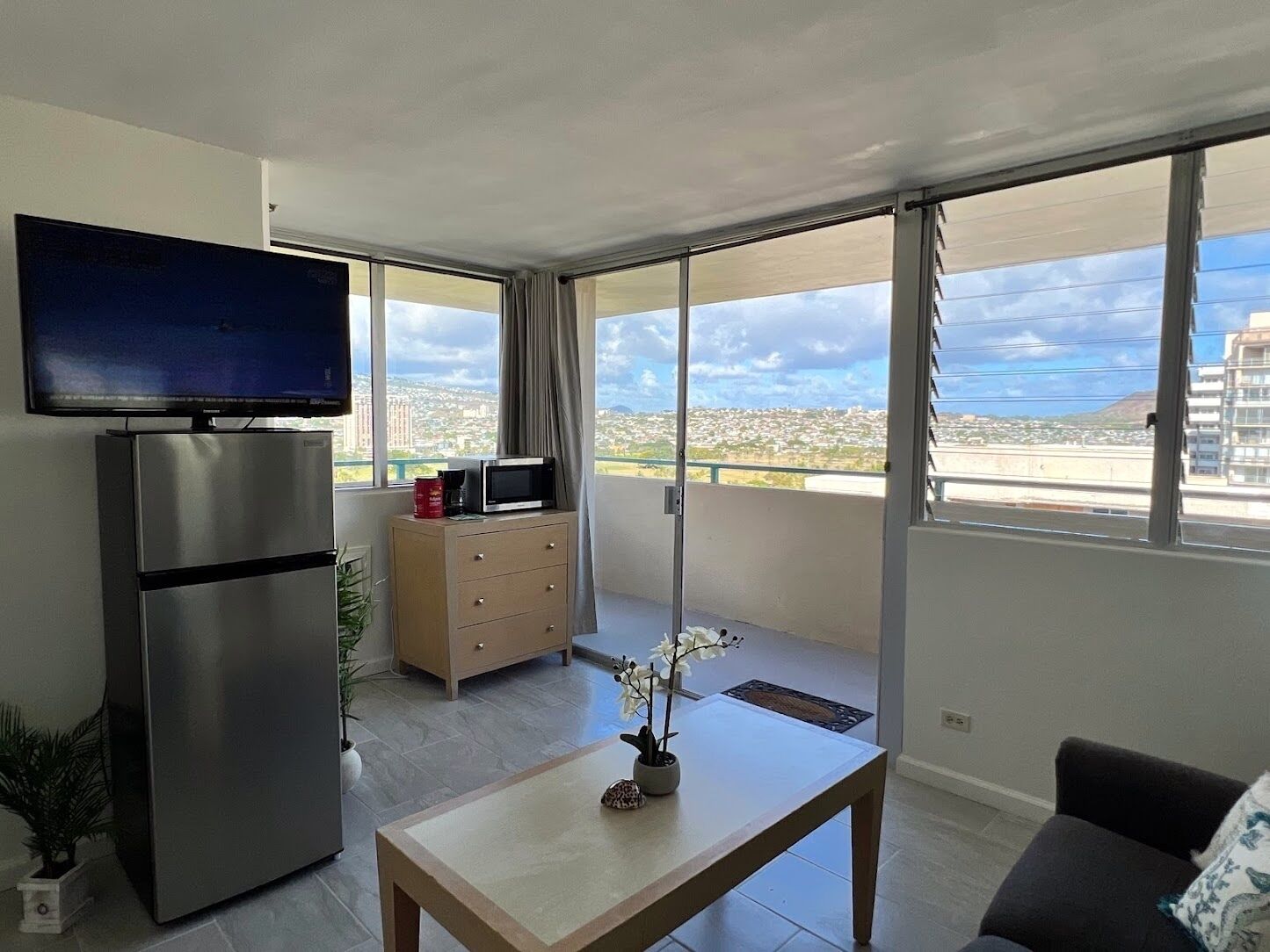 A modern living room with a flat-screen TV, a refrigerator, a microwave, a wooden table, and a sofa. There is a balcony with a city view.