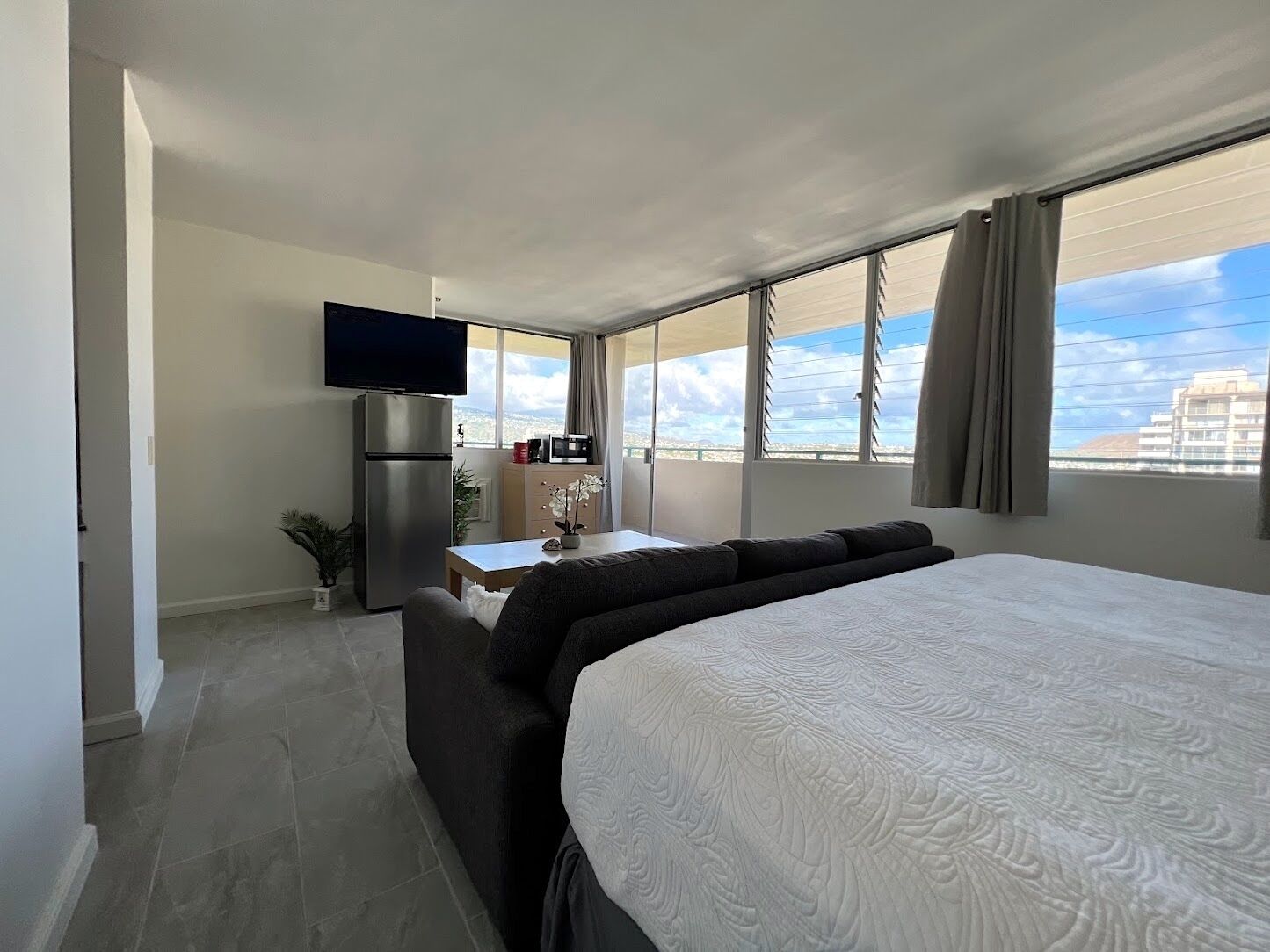 A hotel room with a large bed, a flat-screen TV, a kitchenette, and a view of the city.