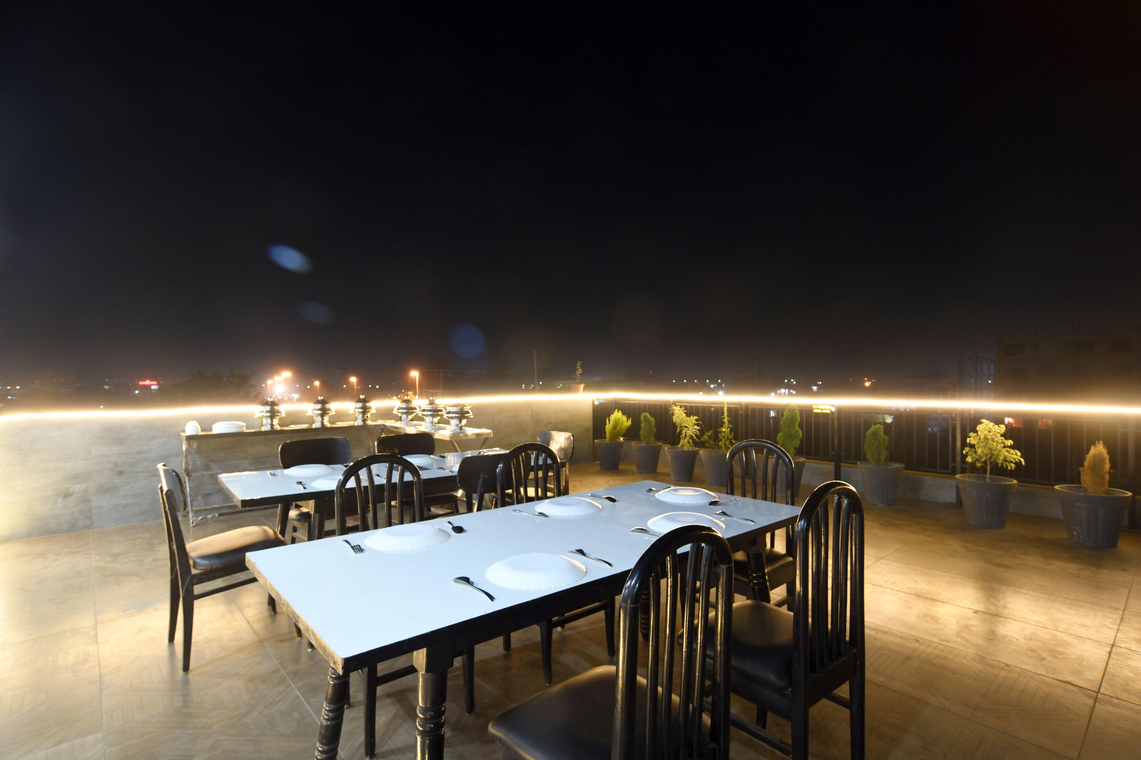 An outdoor dining area with tables and chairs, potted plants, and a view of the city at night.