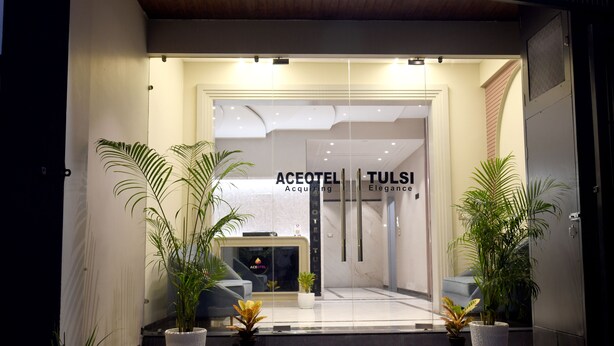 A entrada do Aceotel Tulsi com um design moderno, apresentando um balcão de recepção, plantas em vasos e fachada de vidro.