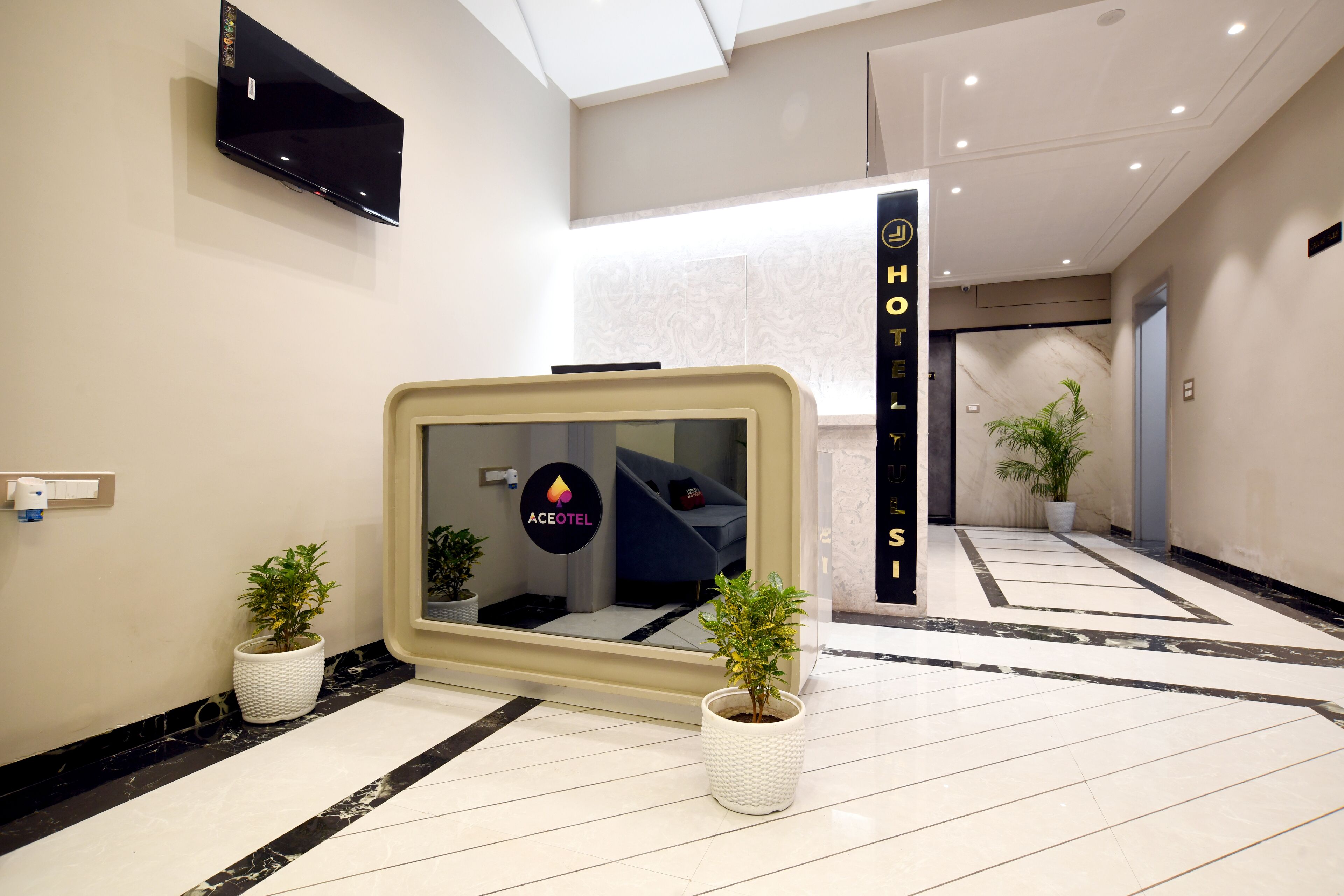 A hotel lobby with a reception desk, a flat-screen TV, potted plants, and a tiled floor.
