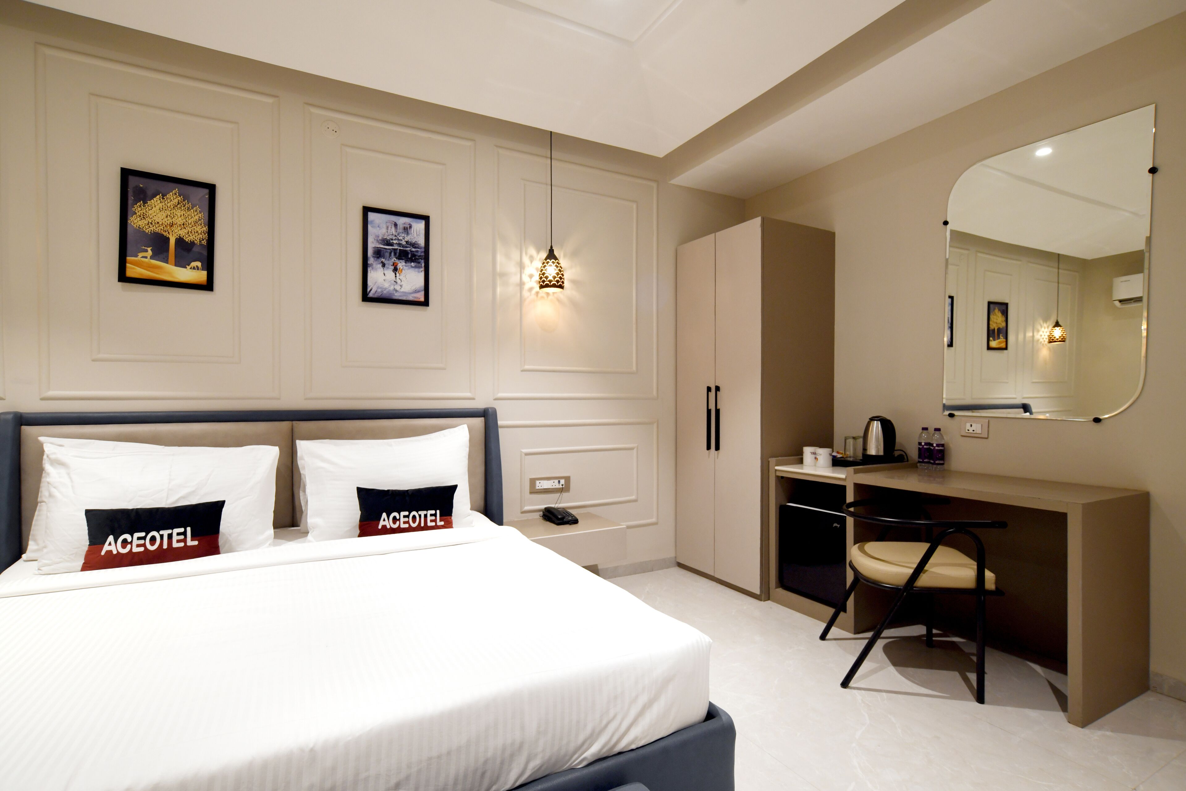 A modern hotel room with a bed, a desk, and a chair. There are framed pictures on the wall and a mirror above the desk.