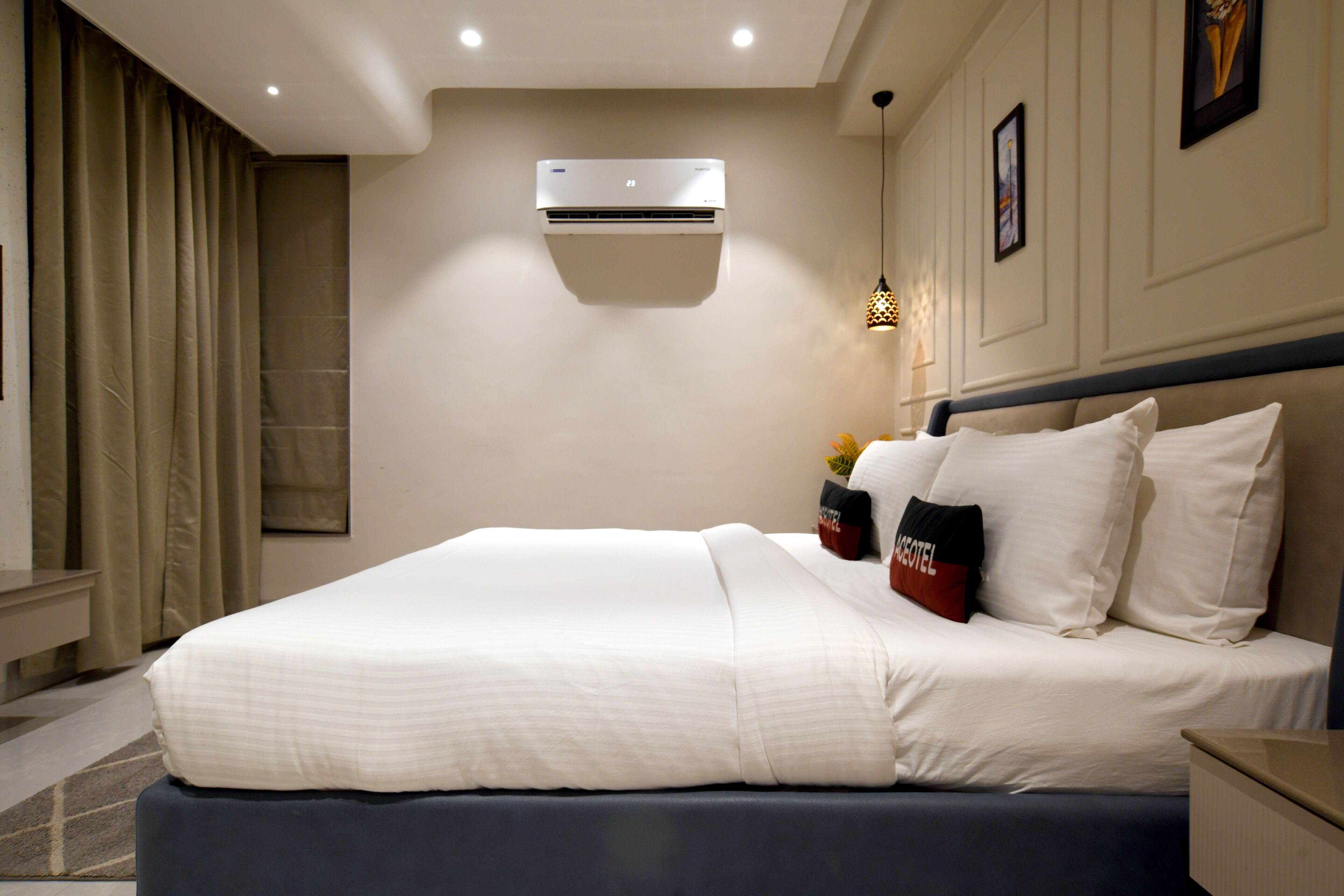 A hotel room with a large bed, a wall-mounted air conditioner, a nightstand, and a lamp.