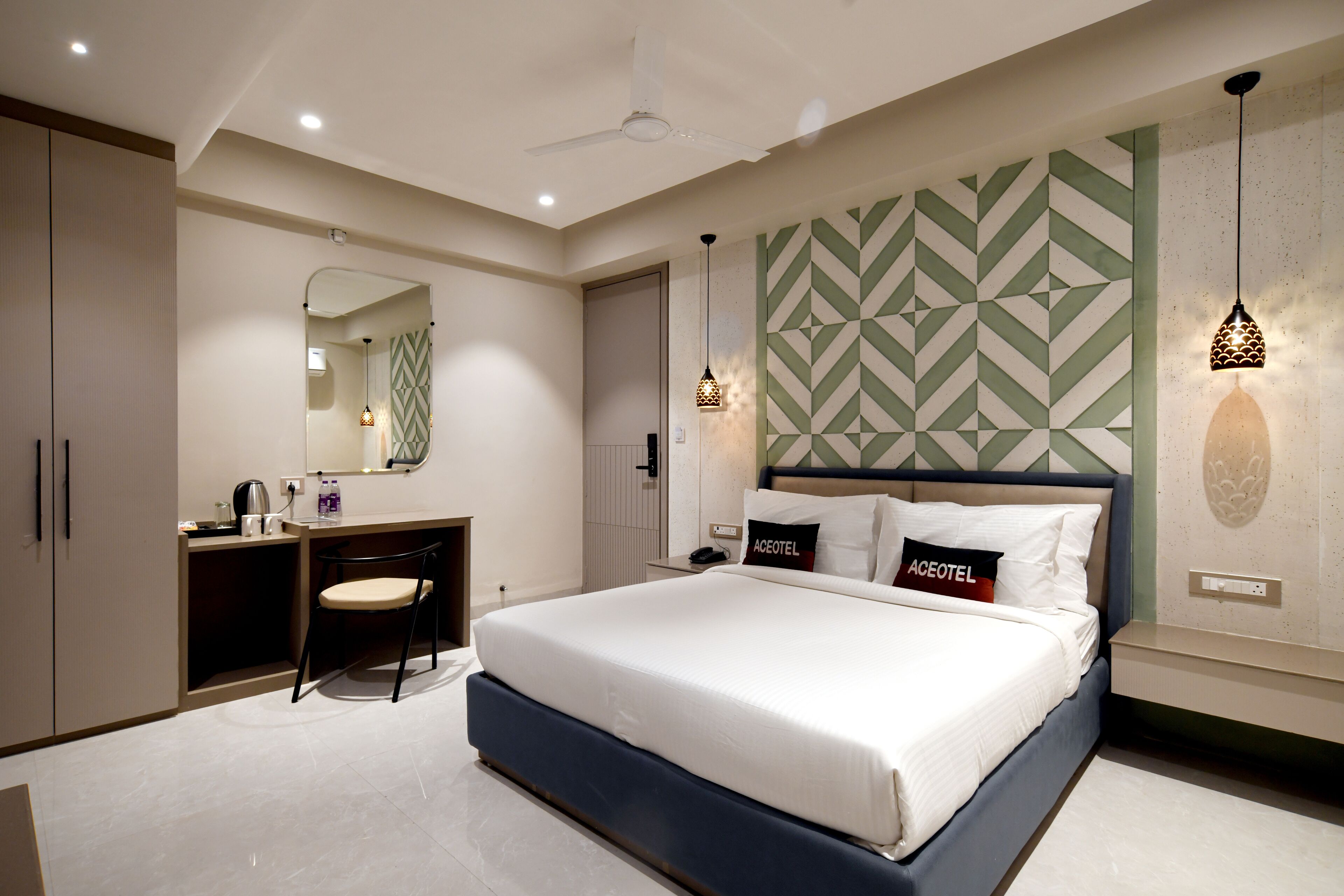 A modern hotel room with a large bed, a desk with a chair, a mirror, and a geometric-patterned wall.