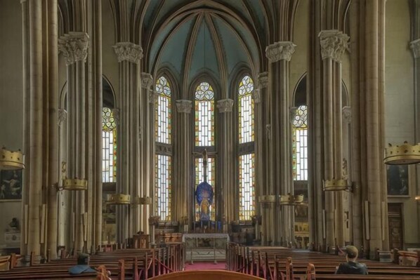 A church interior with high vaulted ceilings, large stained glass windows, and rows of pews.