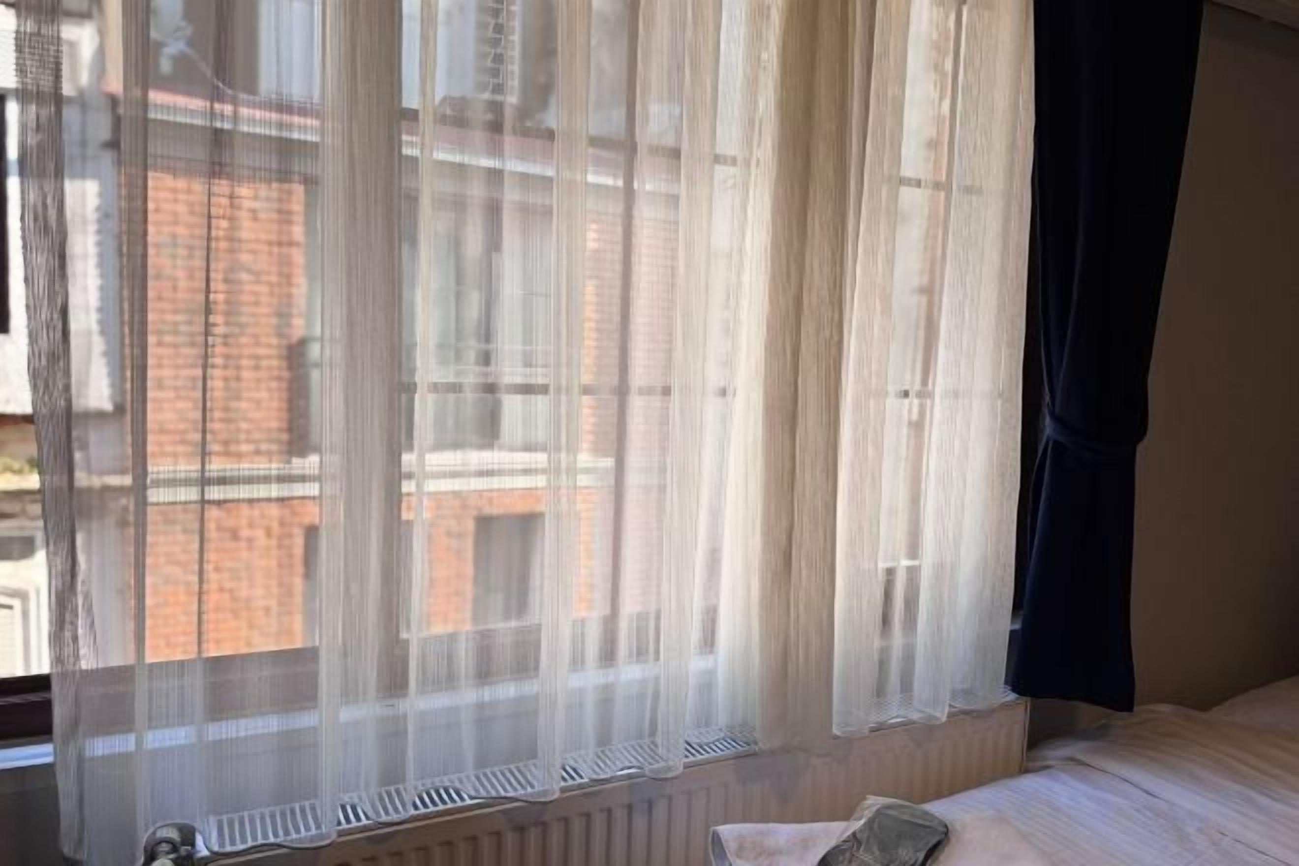 A room with a bed, a window with sheer curtains, and a radiator below the window.