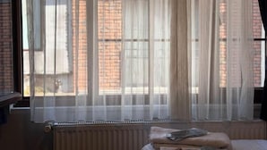 Una habitación con ventana cubierta por cortinas traslúcidas, un radiador debajo y una cama con almohadas y una colcha.