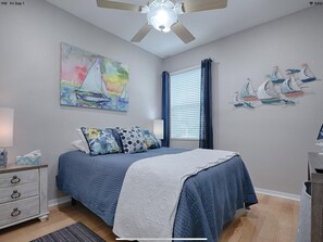 Quarto com cama, ventilador de teto, janela com cortinas, um quadro e decoração náutica na parede.