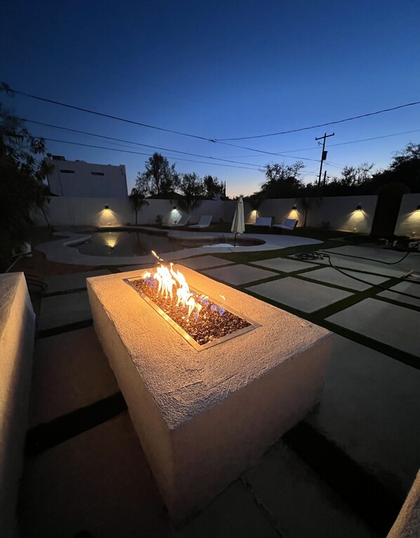 A rectangular fire feature with a fire burning, surrounded by a concrete border, set in a courtyard with lounge chairs and a pool. - Sam Hughes Desert Oasis, Private Pool & Walk to UA (Tucson)