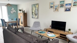 A modern living room with a grey sofa, a TV, and a dining area with a wooden table and chairs. - Maison 4/6 Personnes - Bayeux et Plages du Débarquement (Bayeux)
