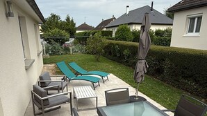 An outdoor patio with a glass-top table and chairs, surrounded by lounge chairs and a swimming pool. - Maison 4/6 Personnes - Bayeux et Plages du Débarquement (Bayeux)