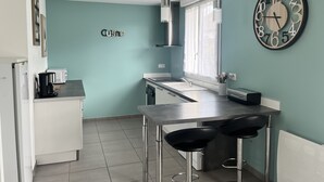 A modern kitchen with a bar area, a clock, and a window with curtains. - Maison 4/6 Personnes - Bayeux et Plages du Débarquement (Bayeux)