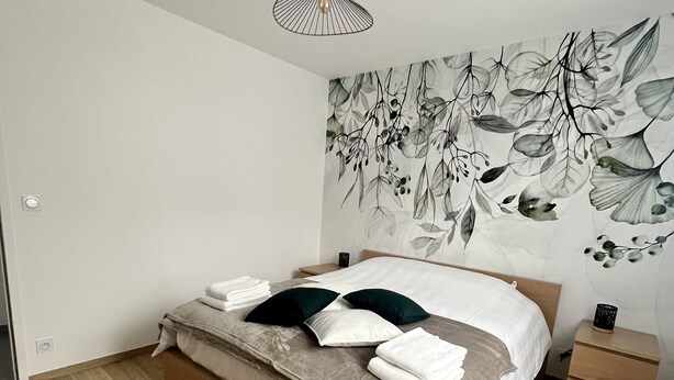A modern bedroom with a wooden floor, a bed with a beige and white duvet, two pillows, a nightstand with a lamp, and a wall with a leaf pattern. - Maison 4/6 Personnes - Bayeux et Plages du Débarquement (Bayeux)