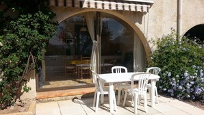 Un patio al aire libre con una mesa y sillas, una puerta de cristal que da a una cocina y un pequeño jardín con flores.