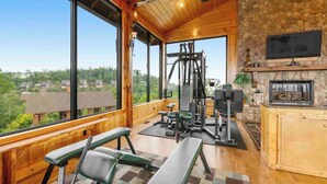 Uma academia com equipamentos de ginástica, lareira e grandes janelas com vista para os arredores.