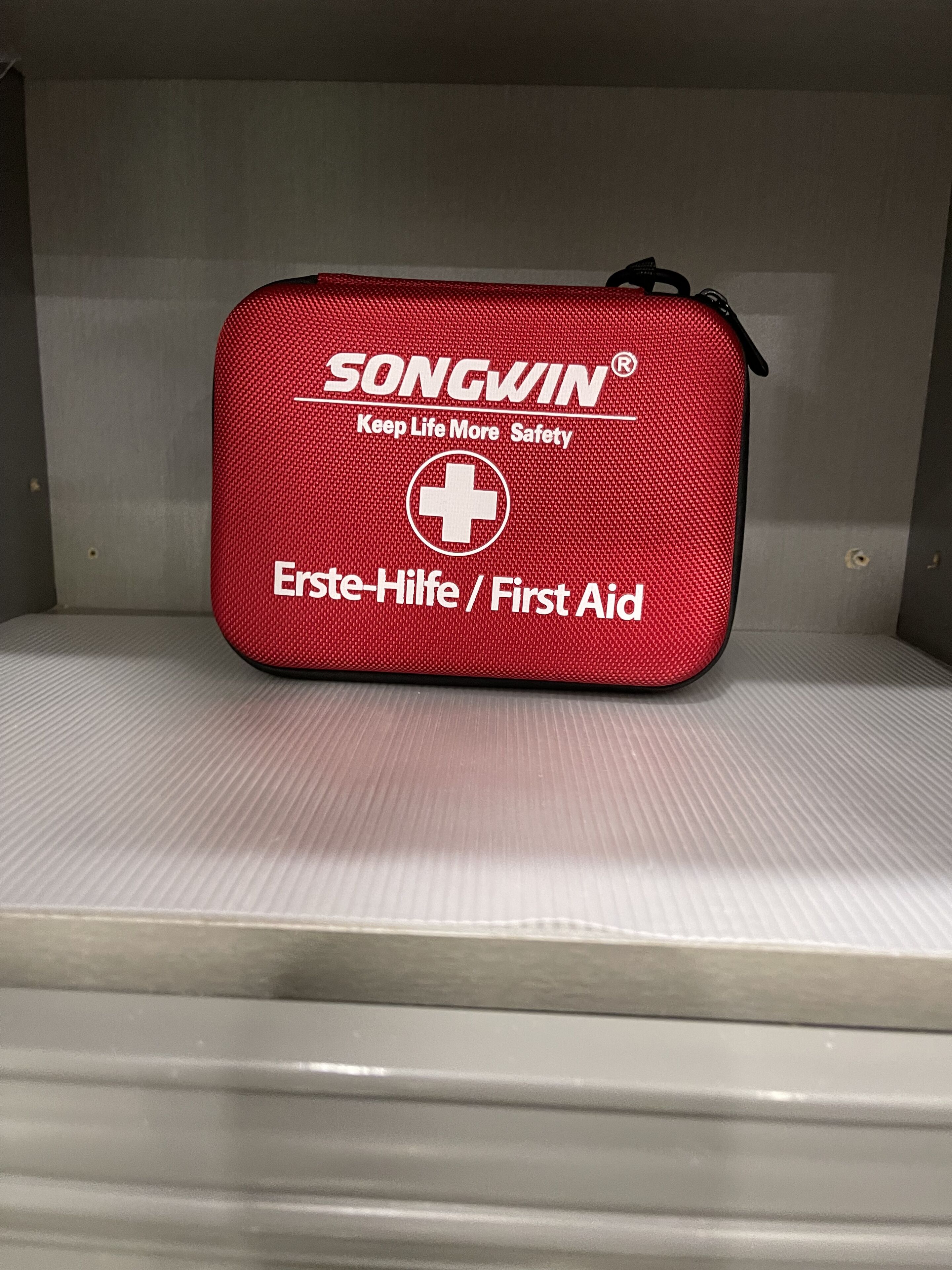 A red first aid kit with the brand name 'SONGWIN' and the text 'Keep Life More Safety' on a shelf.