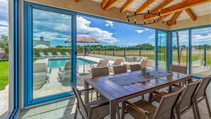 Un pati il·luminat pel sol amb piscina, mobiliari exterior i una gran porta de vidre que dona a una vista del cel i del verdor.