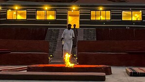 A building with a flat roof and multiple windows, a fire in the foreground, and a desert landscape in the background. - Miral Night Camp (Wadi Rum)
