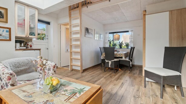 A cozy living room with a sofa, a dining table with chairs, a wooden ladder, and a view of the outside.