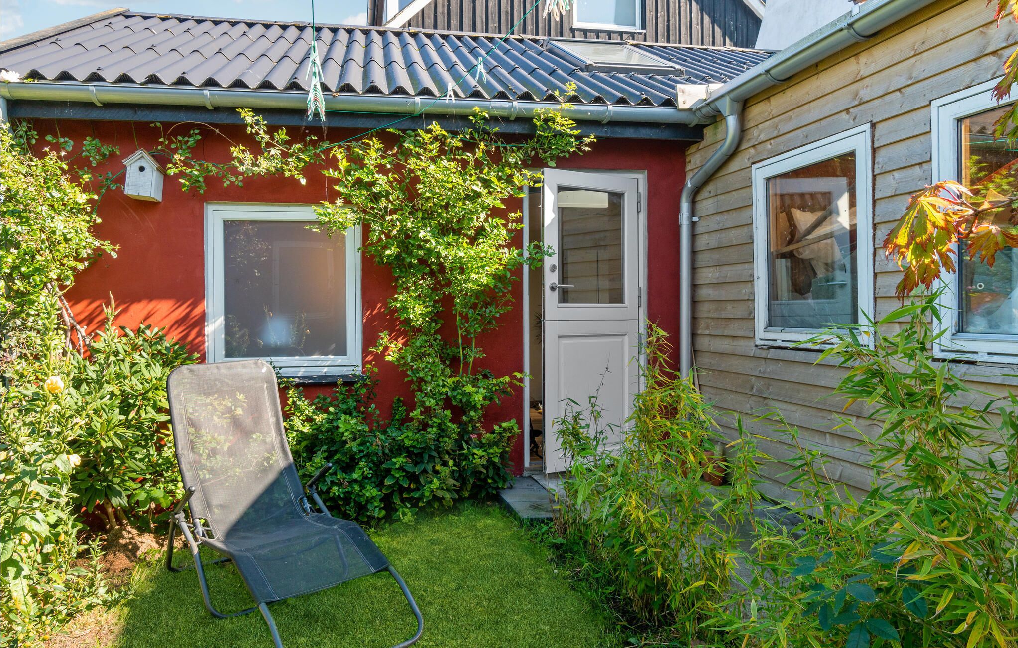 A red house with a white door and window, a small garden with a lawn chair, and a wooden fence.