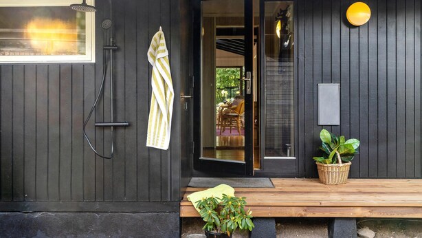 A black wooden building with a glass door, a yellow towel hanging on the door, a wooden bench, and a potted plant. - 3 bedroom awesome home in Gilleleje (Gilleleje)