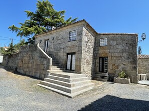 A stone building with a staircase leading to a door, surrounded by gravel and a tree.