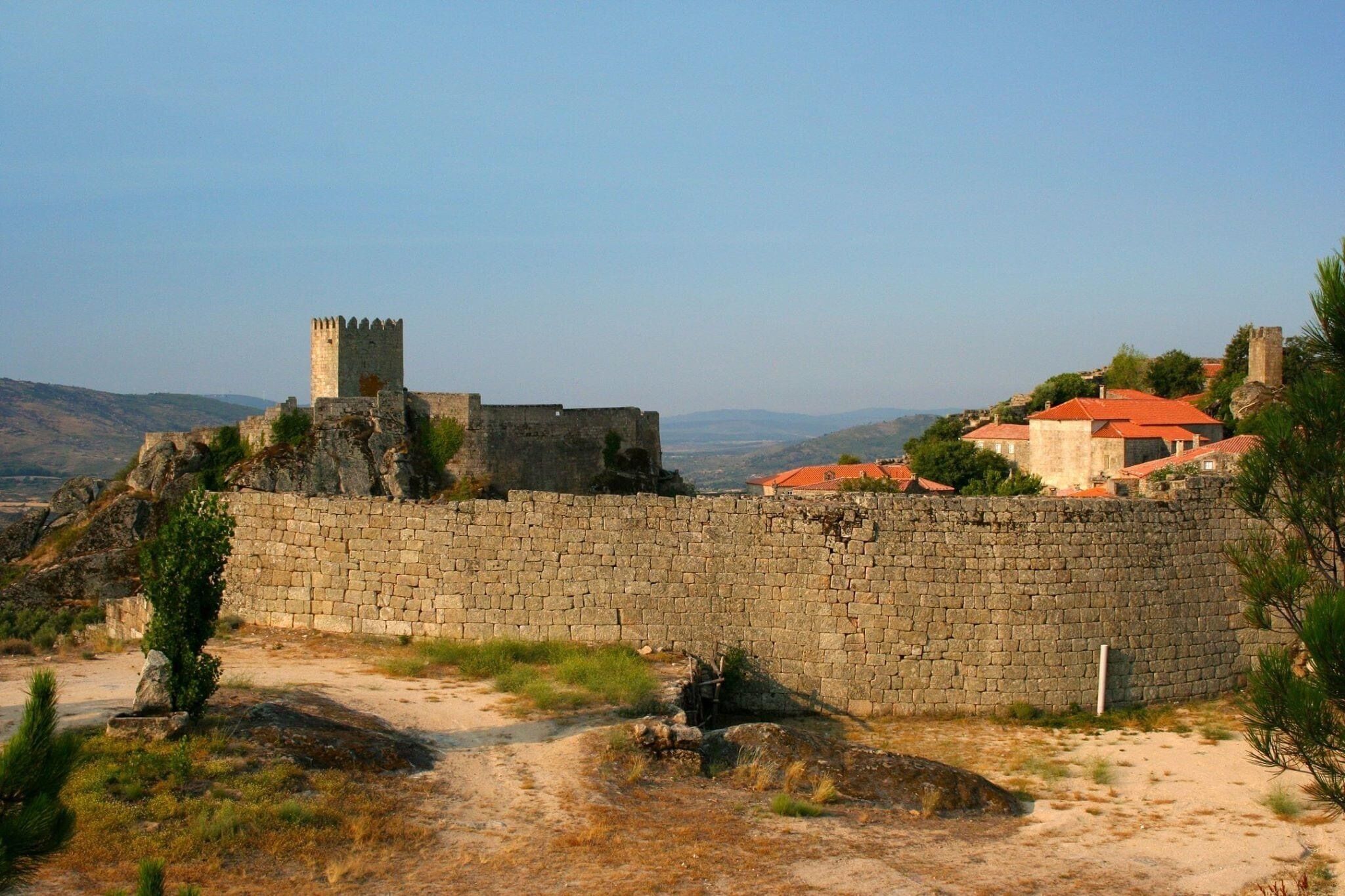 A historic stone wall with a tower, surrounded by a rural landscape and traditional buildings.