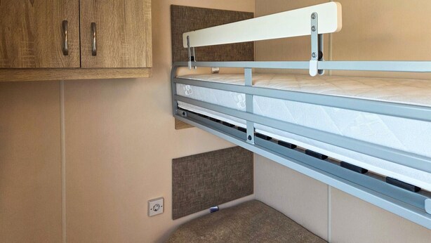Een stapelbed met hoofdbord, bevestigd aan een muur, een klein plankje eronder en een stopcontact in de muur.