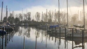 Een jachthaven met aangemeerde boten, stil water en kale bomen op de achtergrond.