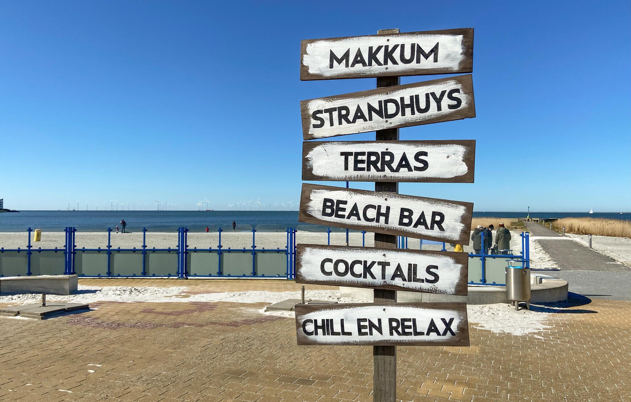 A wooden signpost with multiple destinations listed, including a beach bar and a cocktail area, set against a clear sky and a beach backdrop.