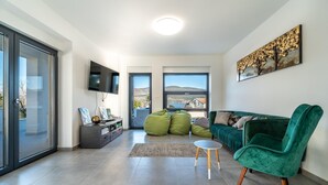 Un salon moderne avec un canapé vert, un téléviseur fixé au mur et une grande fenêtre donnant sur l’extérieur.