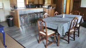 Maalaishenkinen keittiö, jossa on puukaapit, ruokailutila pöydällä ja tuoleilla sekä roskakori.