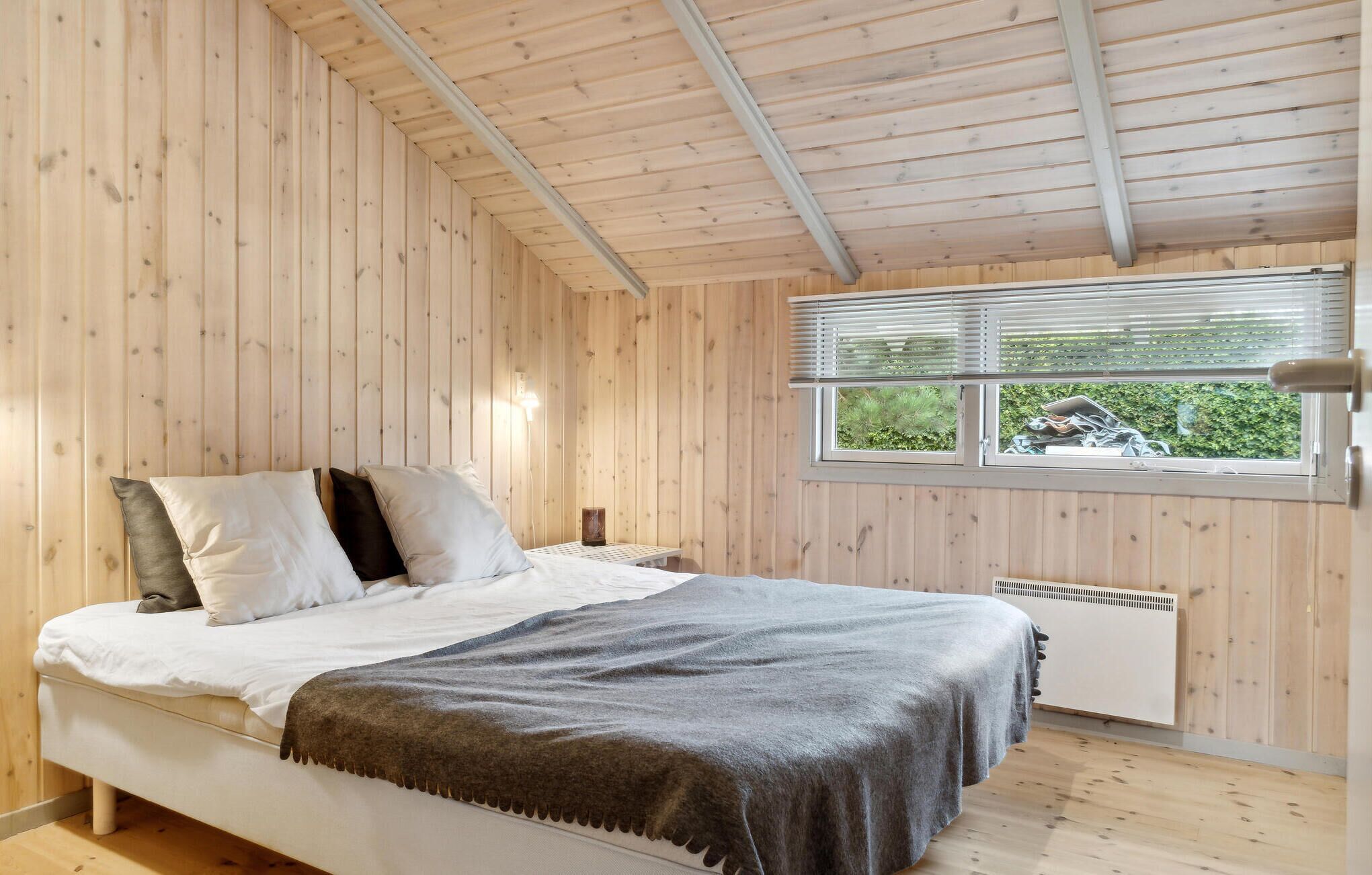 Une chambre à coucher avec un lit, des oreillers, une couverture, une fenêtre avec des stores et un radiateur.