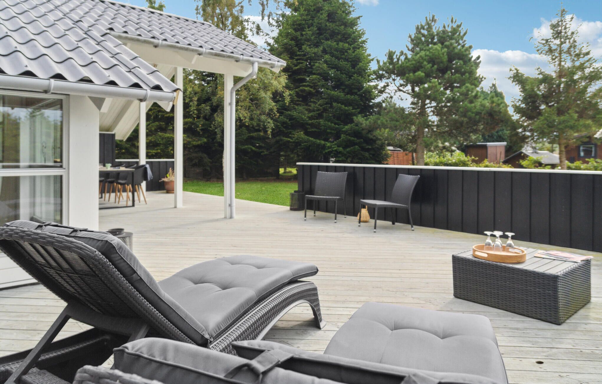 A deck with lounge chairs and a table, a covered seating area, and a view of trees and a house in the background.