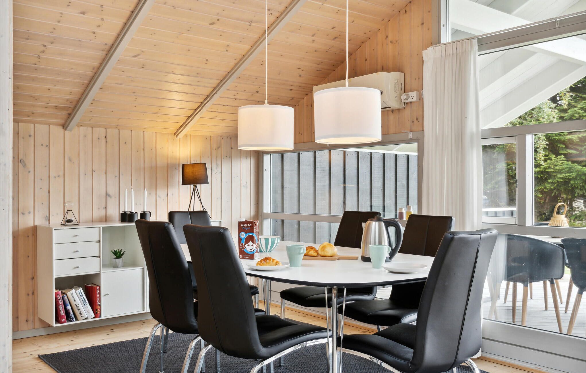 Un espace repas moderne avec une table ronde, des chaises noires et des luminaires suspendus. On y trouve une bibliothèque avec des livres et des objets décoratifs, ainsi qu’une fenêtre avec des rideaux.