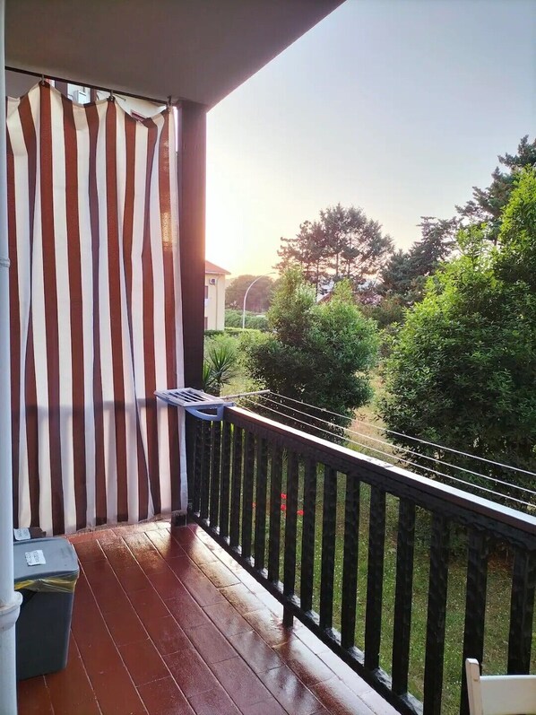 A balcony with a striped curtain, a wooden floor, and a view of a garden and trees.
