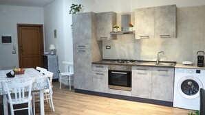 Une cuisine compacte équipée d’une cuisinière, d’un four, d’un évier et d’un lave-linge. On y trouve une table à manger avec des chaises, ainsi qu’une petite table avec une lampe.