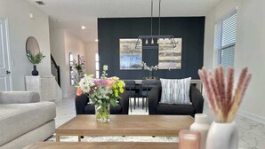 Un salon moderne avec une table basse en bois, un vase de fleurs et un coin repas comprenant un canapé noir et un lustre.