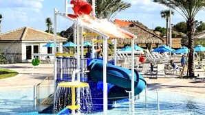 Un parc aquatique avec un toboggan bleu, une aire de jeux avec des jets d’eau aux couleurs jaune et rouge, et des chaises longues placées sous des parasols bleus.