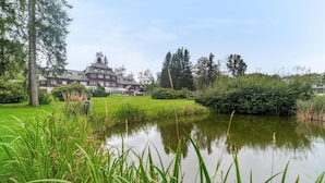 Ein großes Haus mit Teich im Vordergrund, umgeben von üppiger Vegetation und Bäumen.