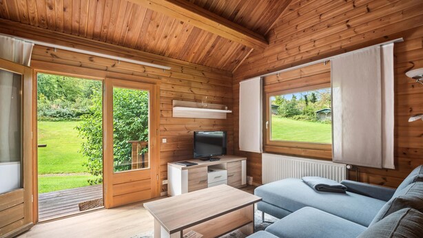 L'intérieur d'un chalet accueillant, avec un plafond en bois, un téléviseur posé sur un meuble en bois, un canapé et une fenêtre donnant sur un paysage verdoyant.