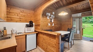 Une cuisine compacte aux murs en bois, avec un réfrigérateur blanc, une machine à café et un four à micro-ondes. On y trouve un coin repas avec une table et des chaises, et une vue sur un jardin grâce à une porte vitrée.